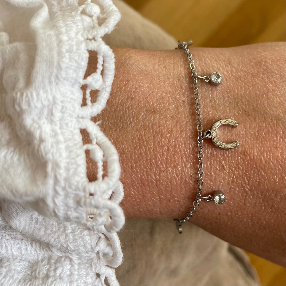 A Ziggy Bracelet with a silver horseshoe charm and two beads adorns a wrist. The wearer’s white, lace-edged sleeve adds elegance against a softly blurred background, highlighting the bracelet’s delicate beauty.