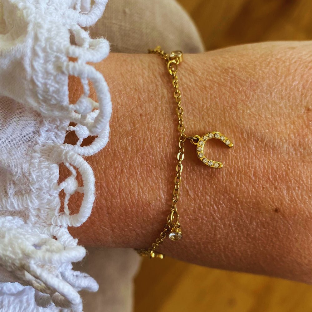 A gold Ziggy Bracelet with a small jewelled horseshoe charm rests on the wrist, styled with a white lacy textured sleeve. The soft, blurred background enhances the delicate look.