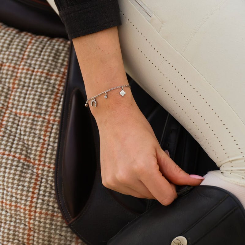 A close-up of a person’s hand resting on a saddle, wearing the Ziggy Bracelet with small pendants. They are dressed in white trousers and a black-sleeved top, seated on textured checked fabric.