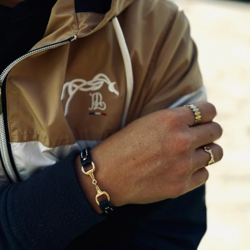 A person wearing a brown and white jacket with a monogram logo crosses their arm, displaying gold rings and a Fano Leather Bracelet with Rhinestones.