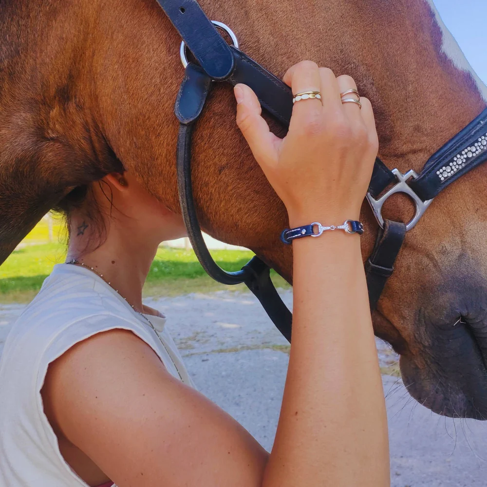 A person gently holds a brown horse’s bridle, pressing their head close to the horse’s neck, showing a close bond. They wear rings and a Fano Leather Bracelet with Rhinestones in the sunny outdoor setting.