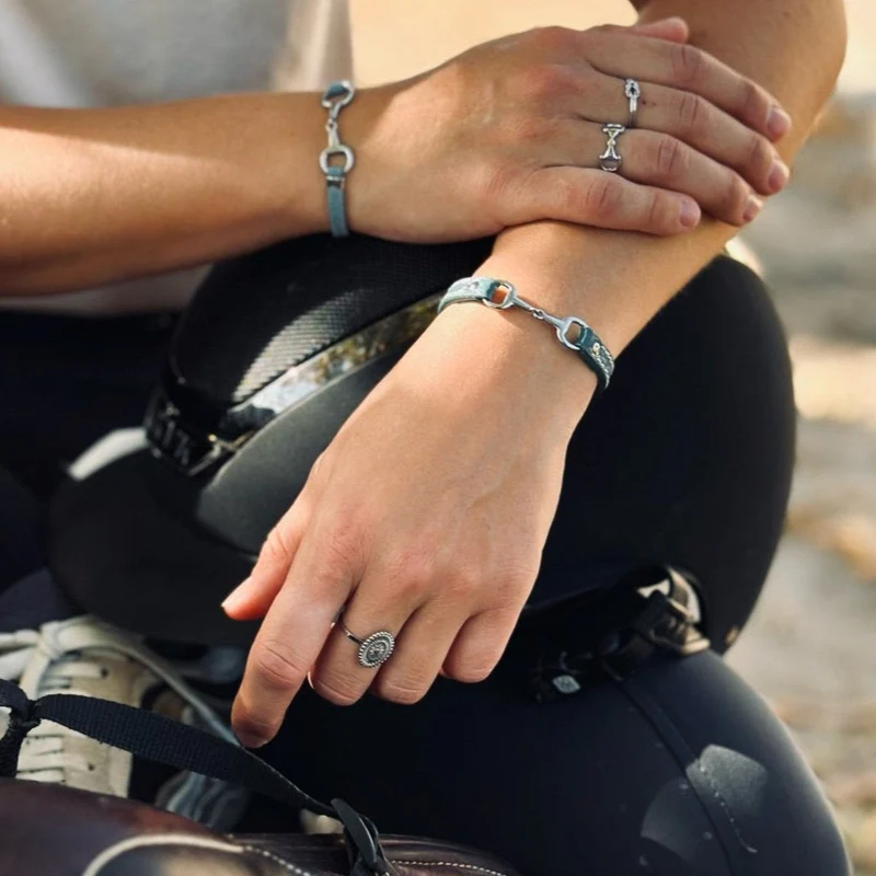 A person rests their hands on a riding helmet, wearing silver rings and a Fano Leather Bracelet with Rhinestones. Dressed in dark trousers, they hold leather reins, adding elegance to the horse riding scene.