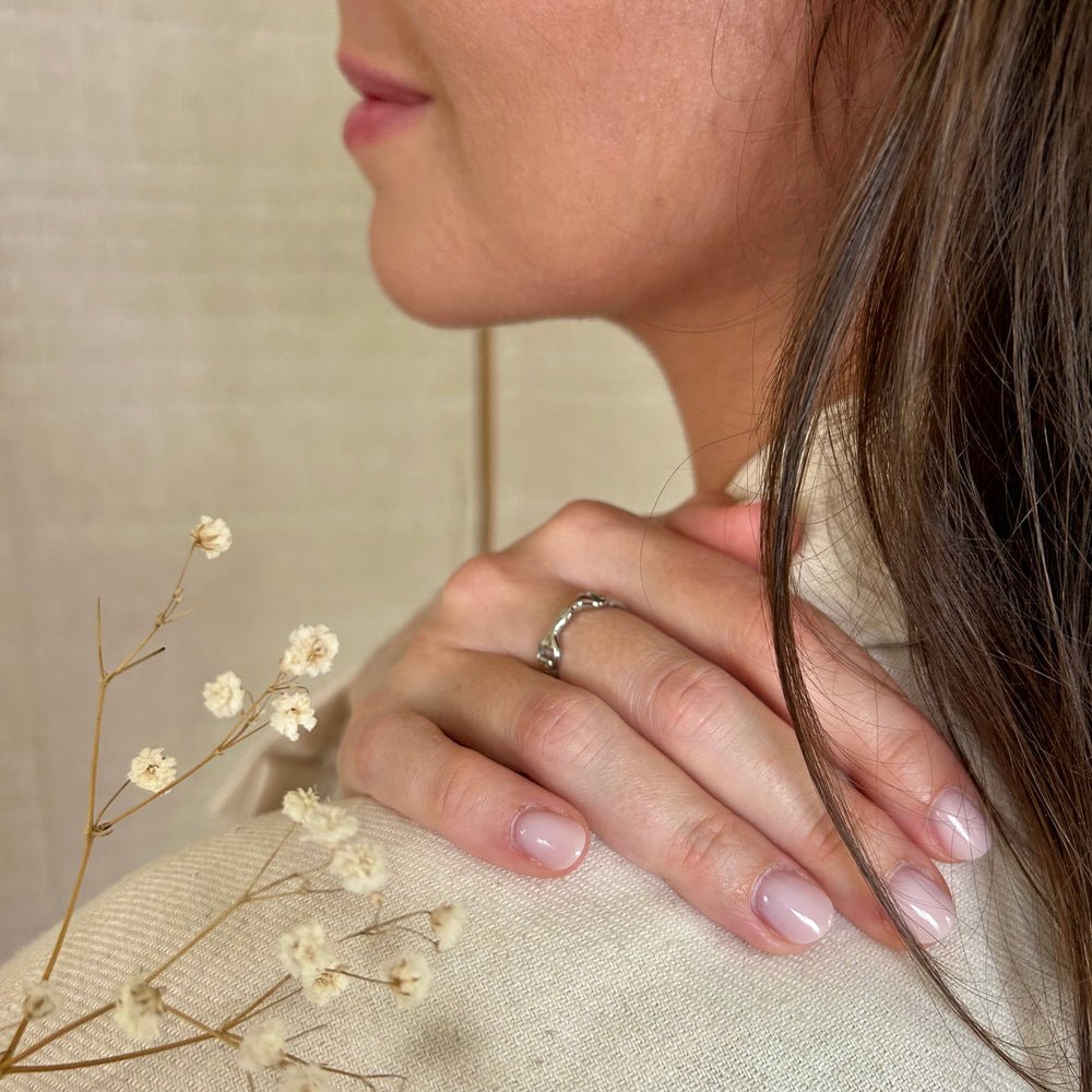 A close-up of a woman’s hand on her shoulder shows light pink manicured nails and the Ana Ring in silver. She wears a beige top, with dried white flowers in the foreground and her face partially visible in profile.