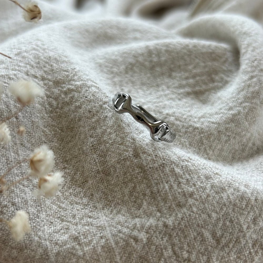 The Ana Ring, a small silver bone-shaped piece, rests on soft textured beige fabric with delicate dried flowers in the foreground.