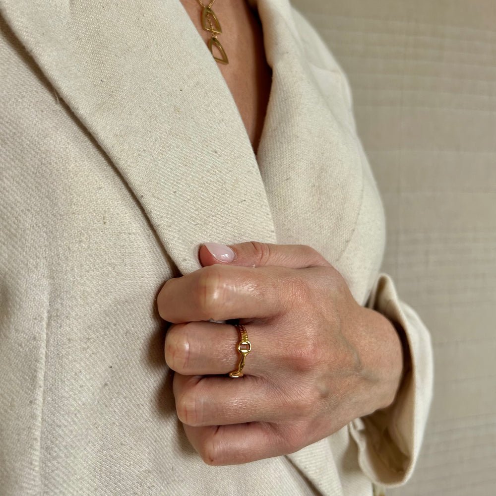 A person wearing a cream-coloured blazer holds the lapel with one hand, displaying the Ana Ring on their finger and a geometric gold necklace. The background is neutral and softly textured.