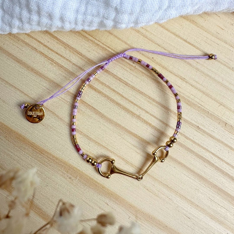 The Sunny Bracelet features pink, gold, and purple beads with a gold horse bit charm on an adjustable pink string, shown on a light wooden surface.