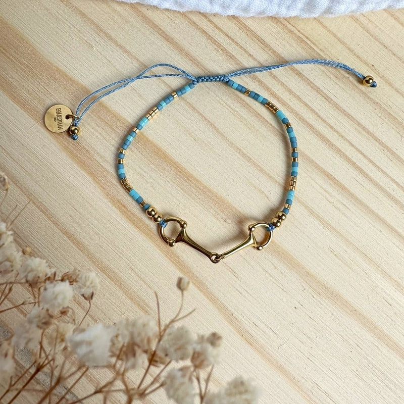 The Sunny Bracelet, adorned with blue and gold beads and a central gold horse bit charm, rests on light wood beside dried white flowers and a round gold tag.