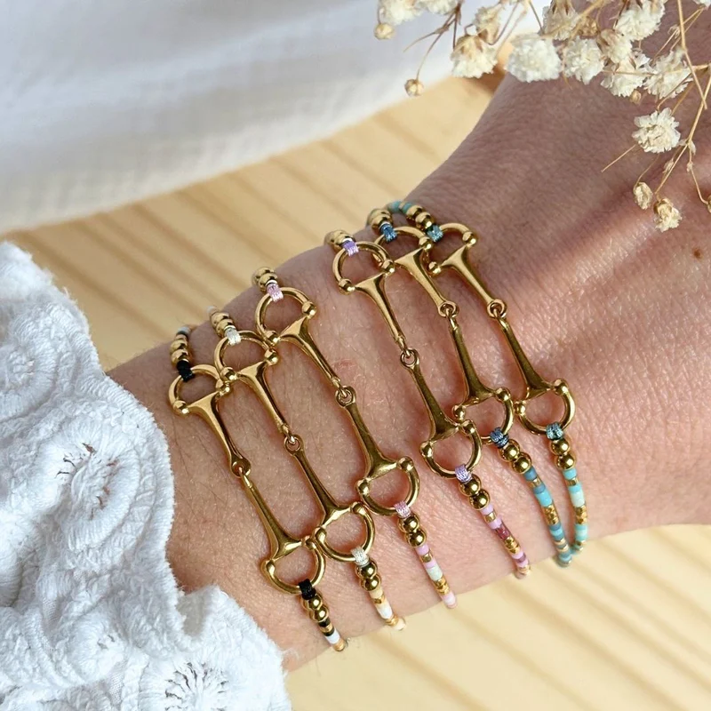 A close-up of a wrist wearing five gold-toned bracelets, including the Sunny Bracelet with colourful beads and equestrian bit charms, beside a white embroidered sleeve and dried flowers on wood.