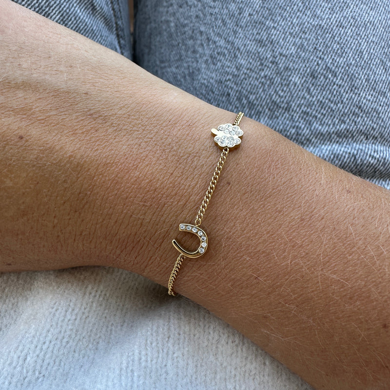 The Willow Bracelet features a gold band with a small horseshoe and clover charm, both set with sparkling clear stones. Shown on the wrist with light denim, it brings luck and elegance to any outfit.