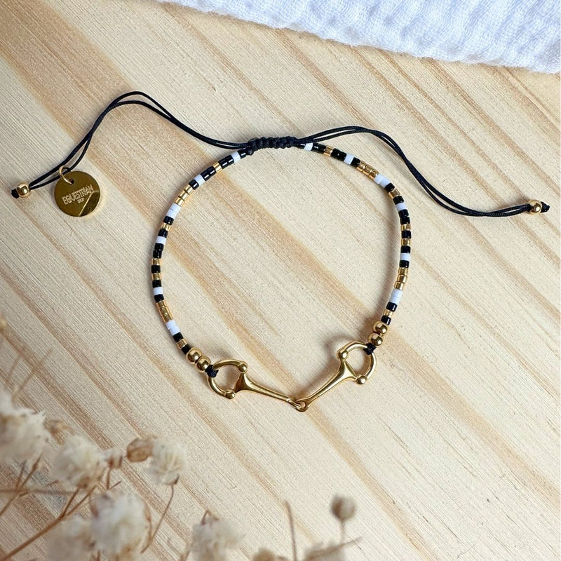 The Sunny Bracelet showcases black, white, and gold beads with a gold horse bit clasp, an adjustable black cord, and a round gold tag, all elegantly presented on a light wooden surface.