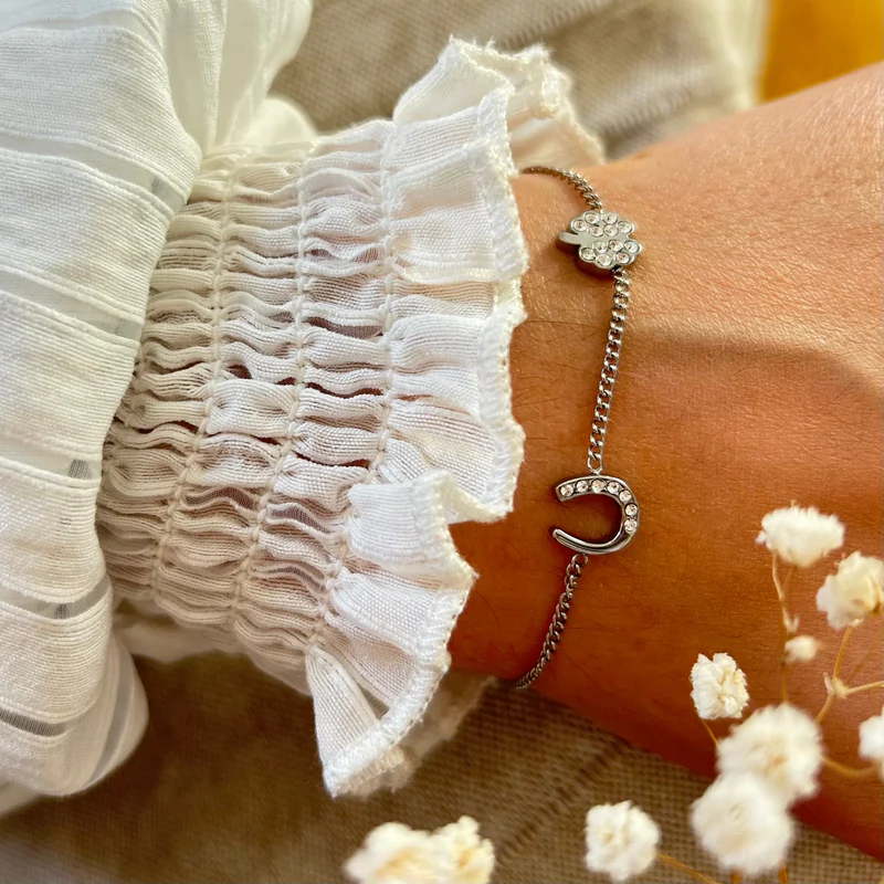 A close-up of the Willow Bracelet, showcasing its silver horseshoe and four-leaf clover charms with crystal details, worn on a wrist in a white ruffled sleeve. Softly blurred white flowers appear in the foreground.