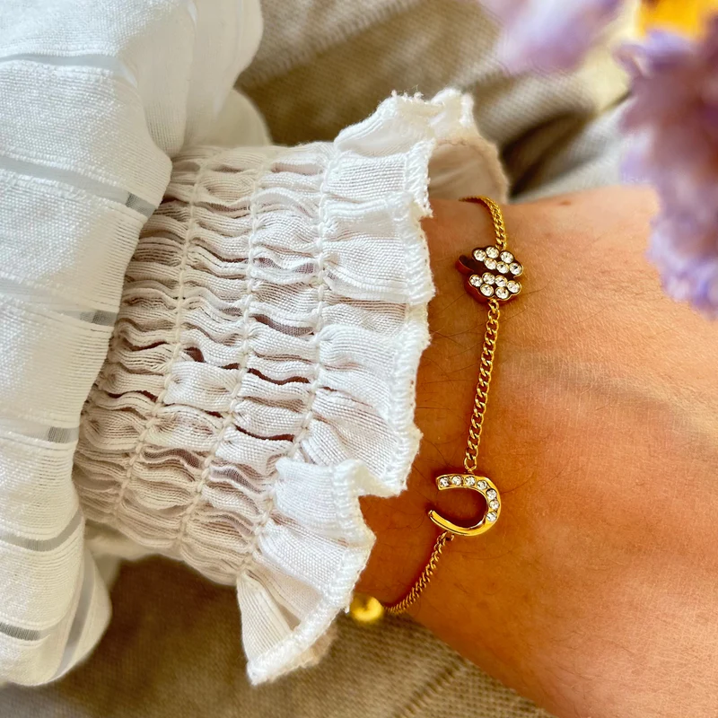 The Willow Bracelet is a gold bracelet with a delicate horseshoe and paw charm set with clear crystals, perfect for wearing on the wrist with a white ruffled blouse.