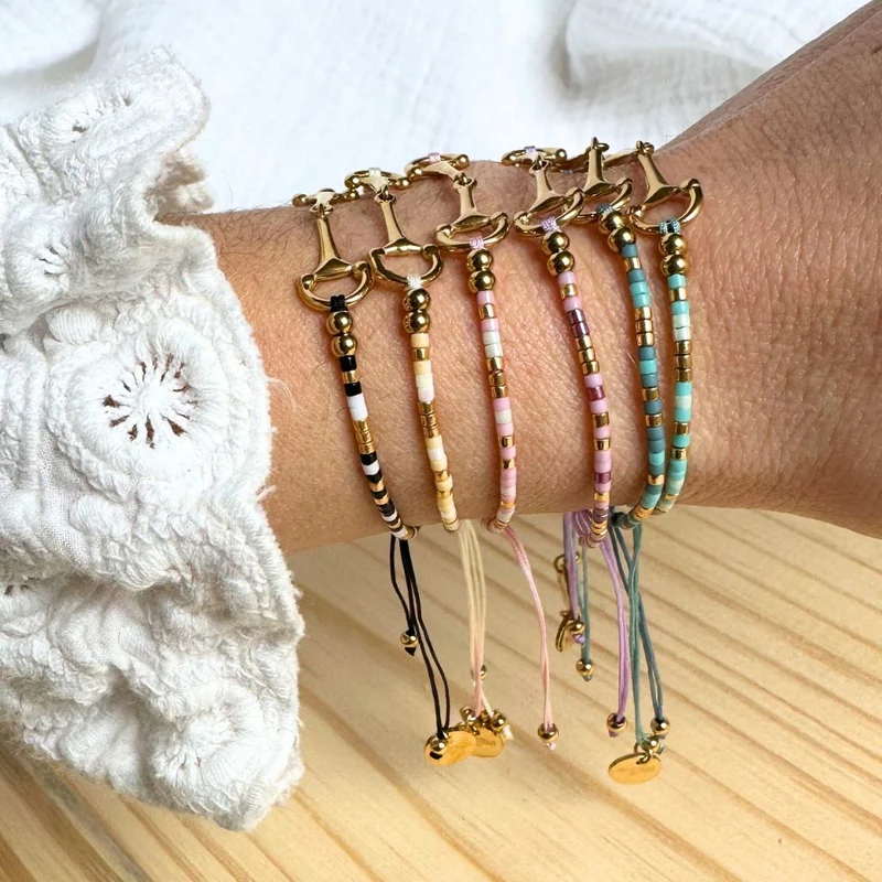 A wrist with six colourful beaded bracelets, including the Sunny Bracelet with gold accents and horse bit charms. Adjustable pastel strings complement a white broderie anglaise-patterned sleeve.