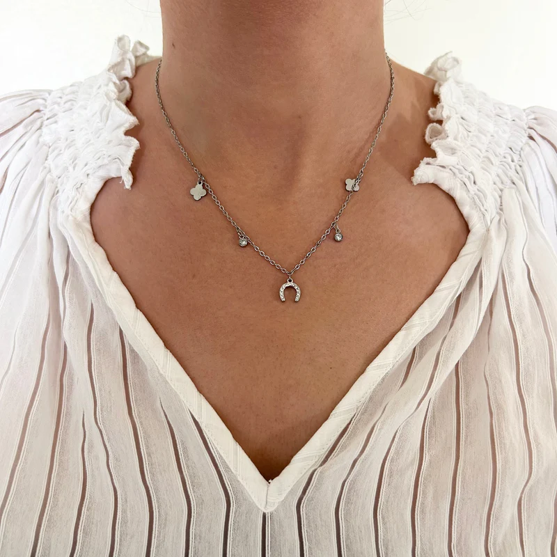A person wearing a white, ruffled pleated blouse and the Ziggy Necklace, which features small charm details, including a horseshoe and butterflies. The photo highlights the Ziggy Necklace on the neck and upper chest.