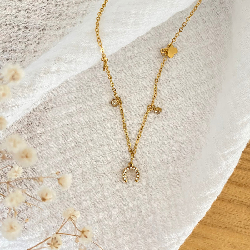 The Ziggy Necklace, featuring a beaded gold crescent moon pendant and small charms, rests on textured white cloth beside dried flowers atop a wooden surface.