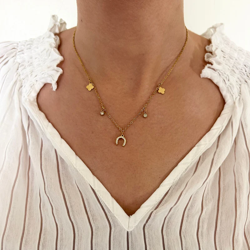A person wearing a white pleated blouse with a ruffled collar and the Ziggy Necklace, which has a gold horseshoe pendant and small charm details.