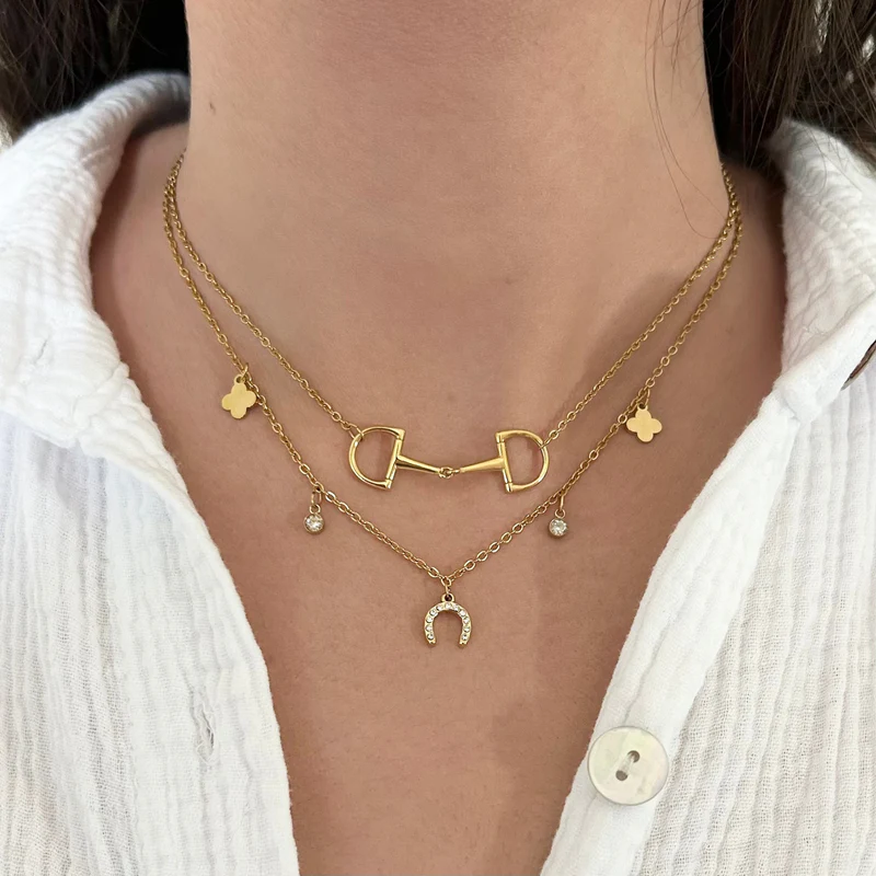 A person wearing a white textured shirt and three layered gold necklaces, one being the Ziggy Necklace with horse bit, clovers, gems, and a horseshoe charms.