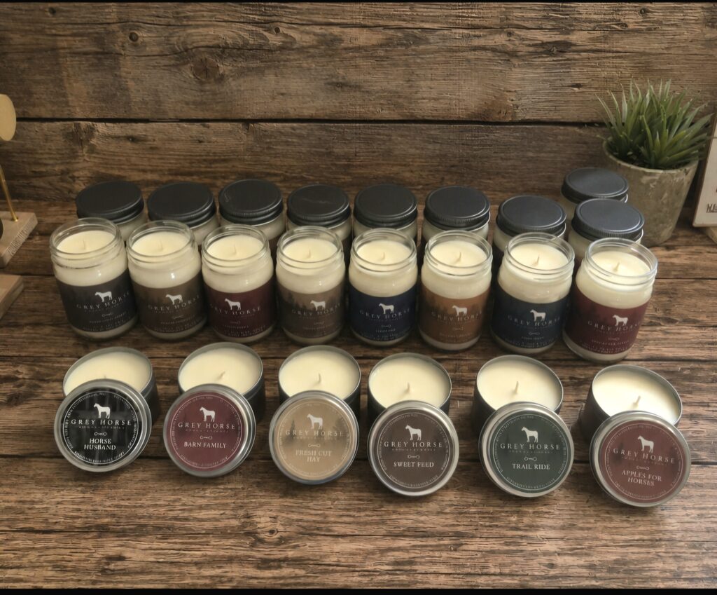 Twelve Grey Horse Candle jar candles with black lids, each featuring a different scent label, are arranged in three rows on a wooden surface. In front, five open Grey Horse Candle jars sit without lids, with a small plant in the background.