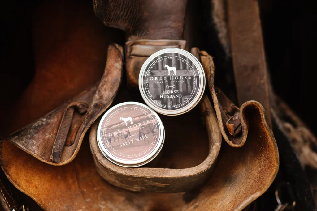Two Grey Horse Candle Tins—Horse Husband and Salty Mare—sit on a worn leather saddle, highlighting the equestrian-themed Grey Horse Candle Tins.