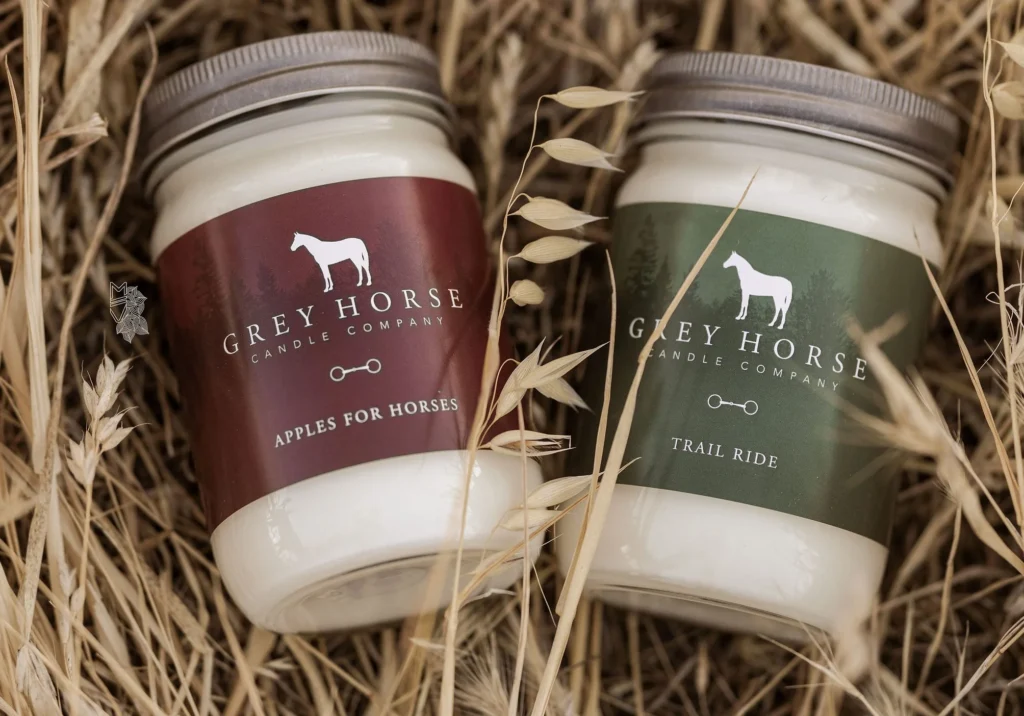 Two Grey Horse Candle jars rest in dry grass—one with a red Apples for Horses label, the other with a green Trail Ride label. Both jars feature the signature white horse silhouette on front.