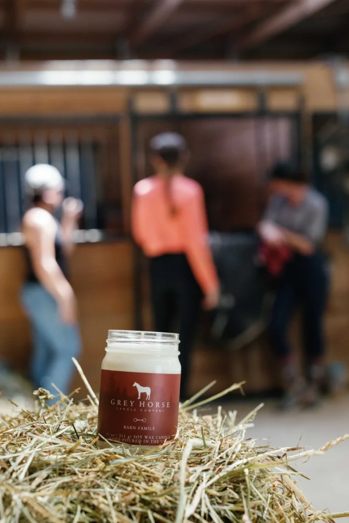 A Grey Horse Candle rests on hay inside a stable, with three people and a horse blurred in the background near wooden stalls, evoking the rustic charm of Grey Horse Candle.