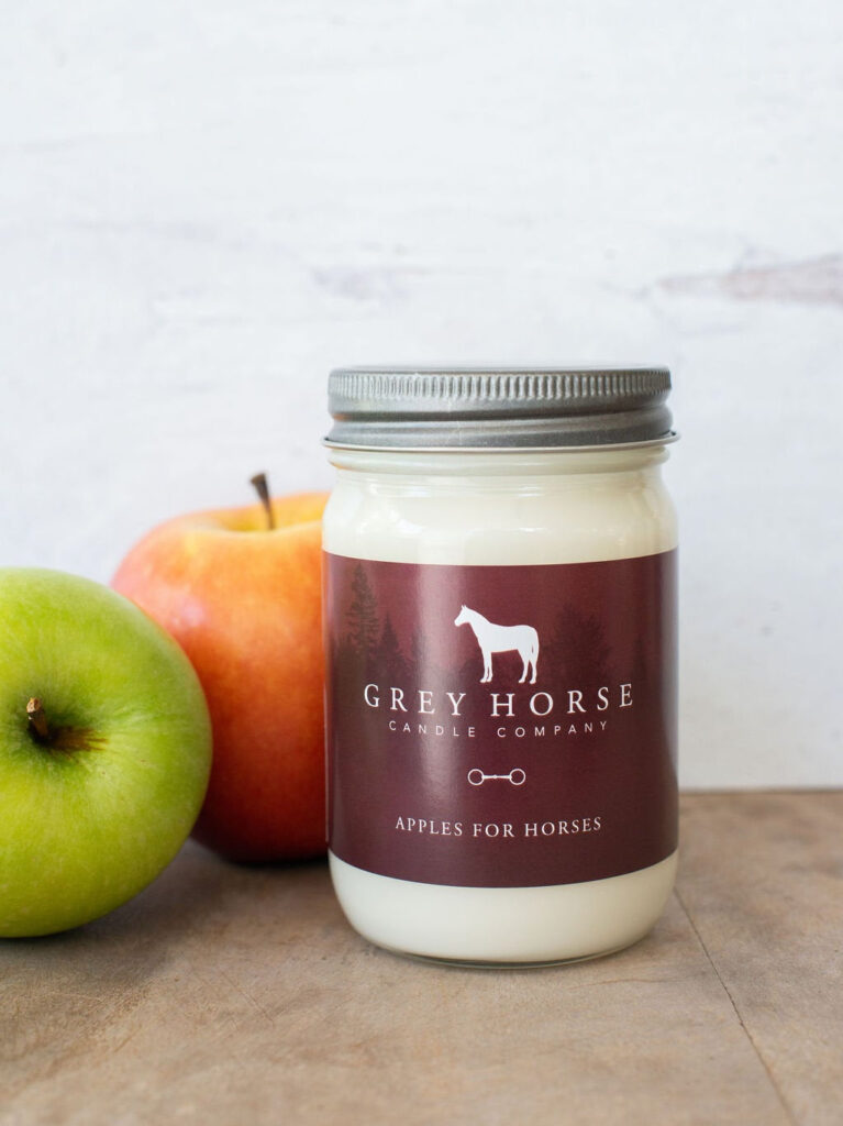 A jar candle labelled Grey Horse Candle Company: Apples for Horses sits on a wooden surface next to a green apple and a red apple, against a light background.