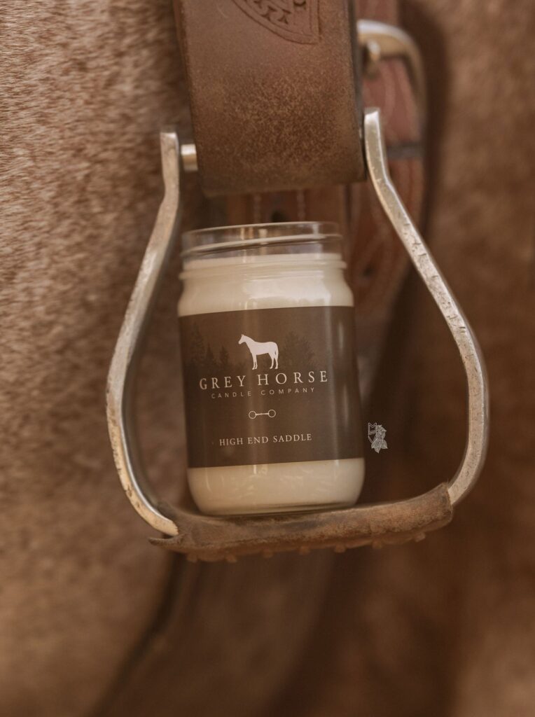 A High-End Saddle Jar candle from Grey Horse Candle Company rests inside a metal stirrup attached to a brown leather saddle on a horse.