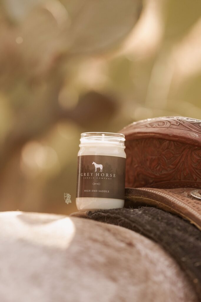 A High-End Saddle Jar from Grey Horse Candle Co. rests on a detailed brown leather saddle, with a softly blurred backdrop and part of a horse’s back visible in front.