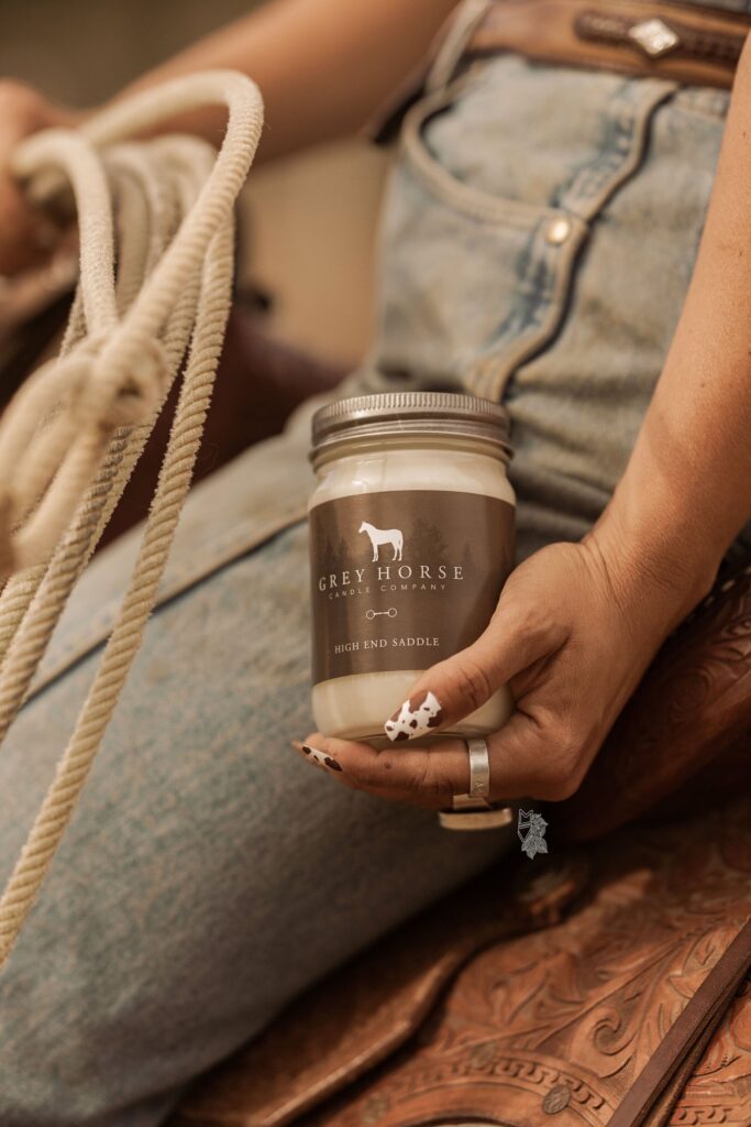 A person in jeans, with painted nails and rings, sits on a saddle holding a rope and the High End Saddle Jar from Grey Horse Candle Co.