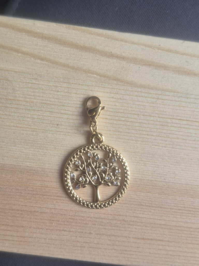 The Lucky Charm Keyring features a gold round pendant with a delicate tree design adorned with small crystals and attached to a lobster clasp, all resting on a light wooden surface—an elegant accessory that serves as both a lucky charm and a beautiful keyring.