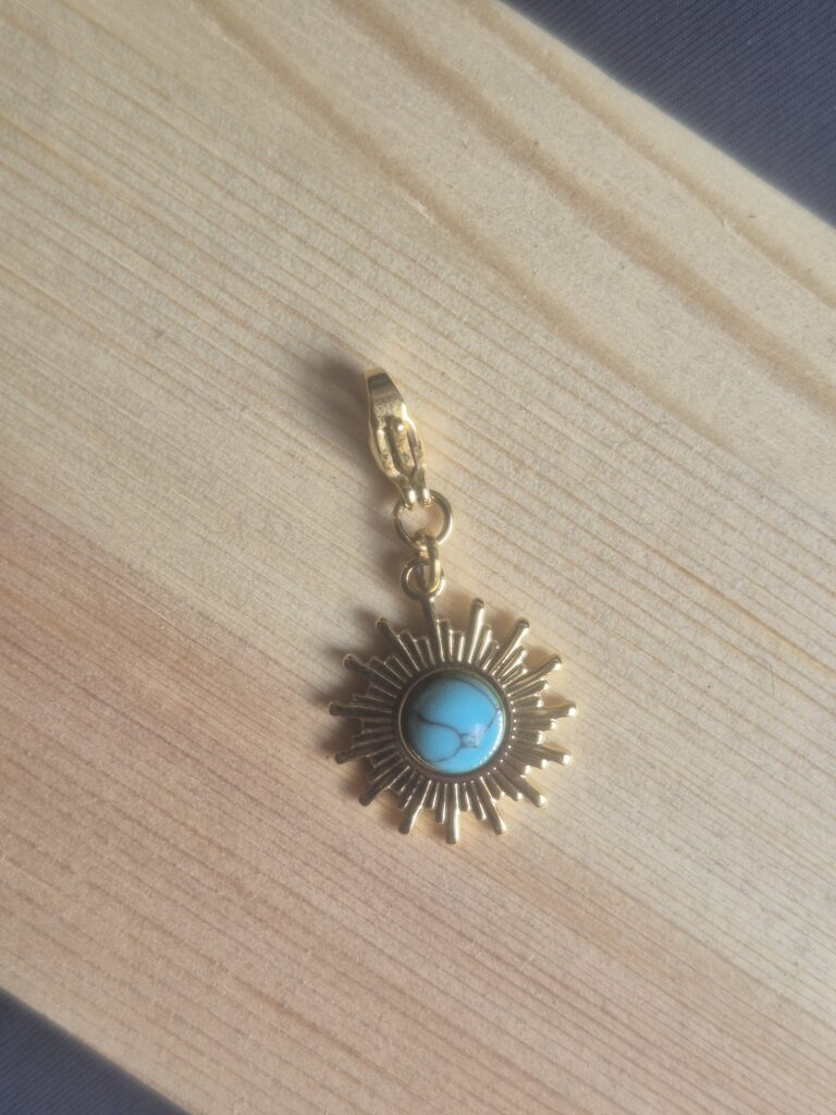 A gold sun-shaped Lucky Charm Keyring with a turquoise centre rests on light-coloured wooden planks, standing out as a unique accessory against the grey backdrop.