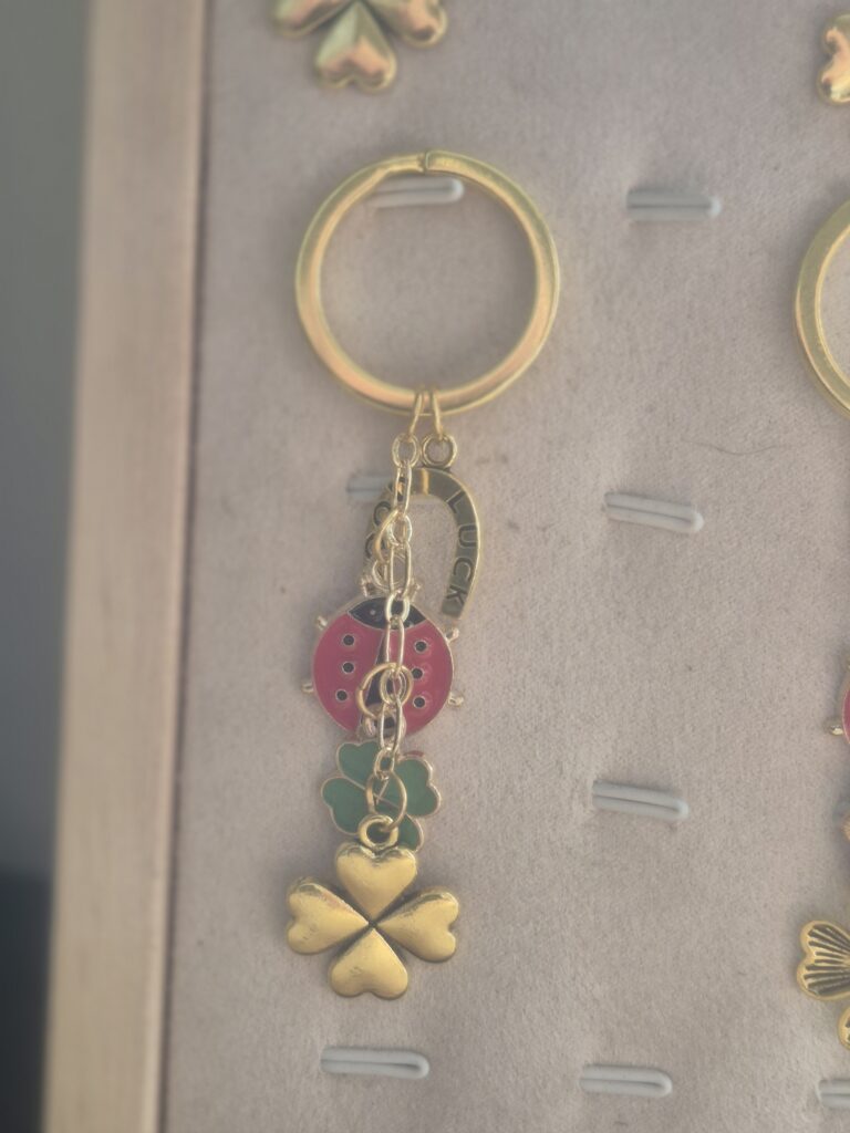 The Lucky Charm Keyring features a gold keyring adorned with hanging charms: a red ladybird, two green shamrocks, a horseshoe, and a gold four-leaf clover, all displayed on a beige background—the perfect lucky charm accessory.