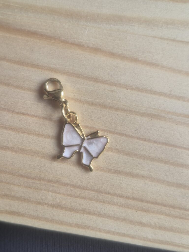 The Lucky Charm Keyring, featuring a gold butterfly charm with white enamel wings and a clasp, lies on a light wooden surface—making it a delightful token of luck or a stylish accessory for your keys.