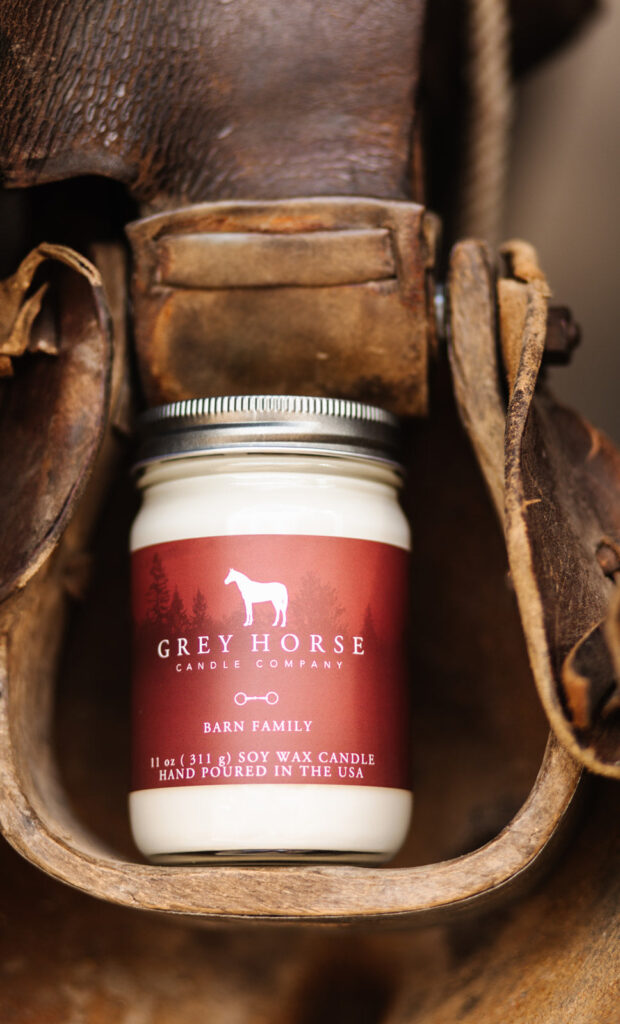 A Barn Family Jar candle from Grey Horse Candle Company, hand-poured with soy wax, sits in a worn leather saddle. The candle has a silver lid and a red label adorned with a white horse graphic.