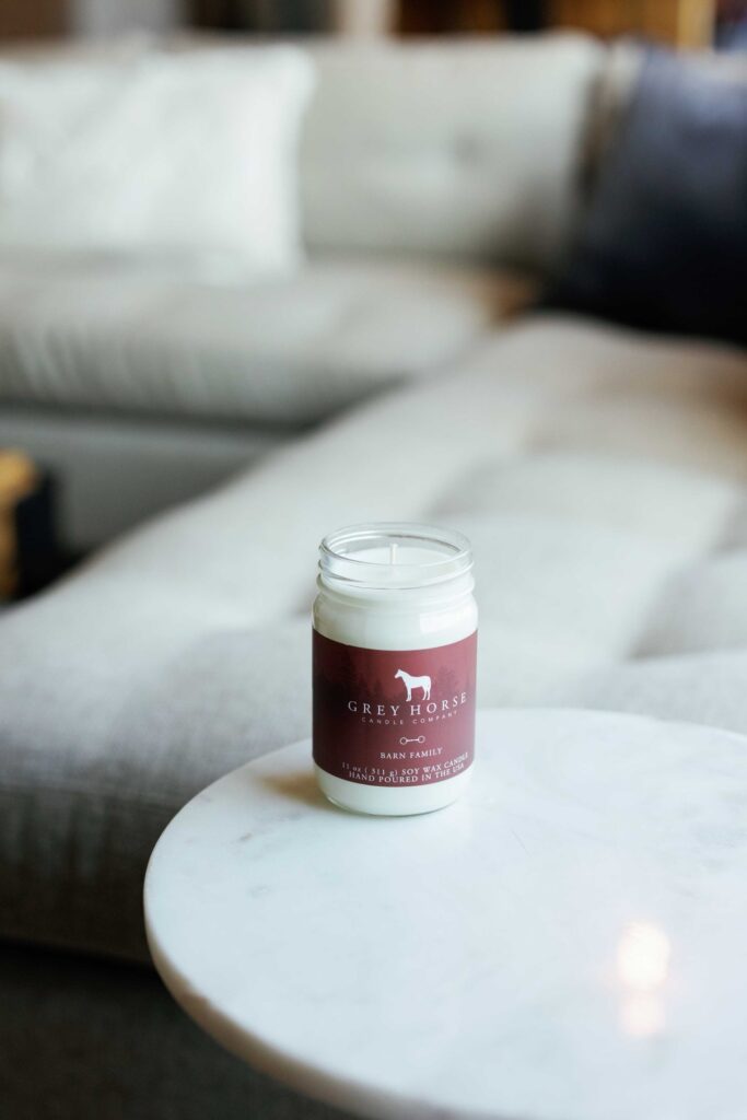 A white Barn Family Jar candle in a glass container sits on a round white marble table in front of a light-coloured sofa with cushions, creating a cosy, softly lit living room reminiscent of warm barn gatherings and family comforts.