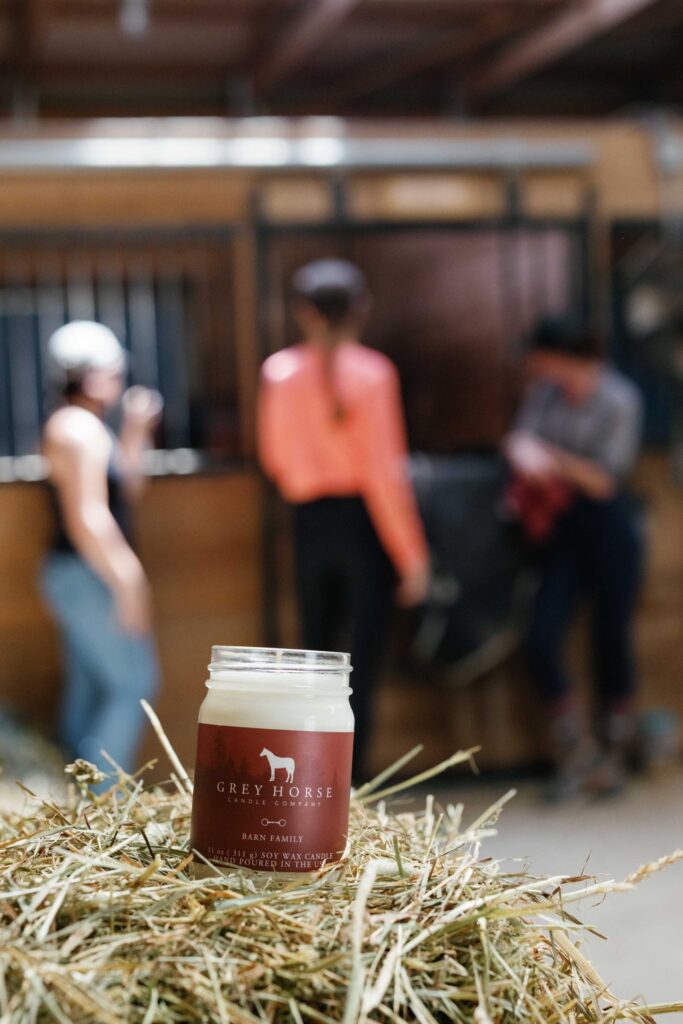 A Barn Family Jar candle sits on a pile of hay in a stable, with three blurred people and a horse in the background, evoking the warmth of barn family gatherings.