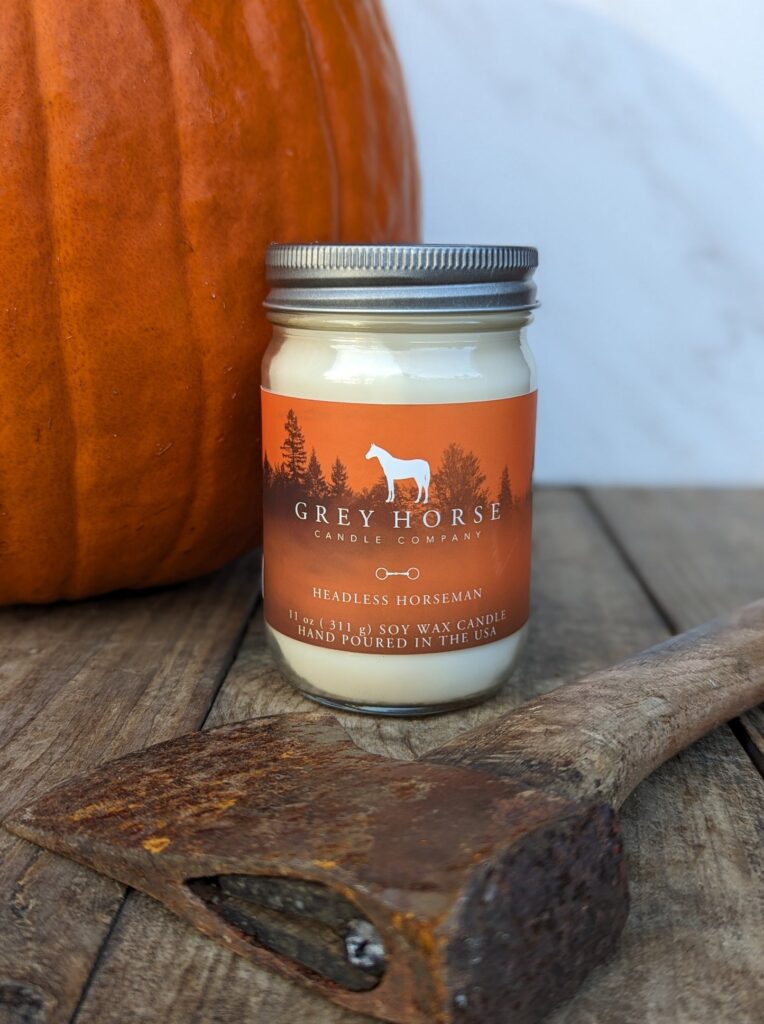 A Headless Horseman Jar rests on a wooden surface beside an axe head and an orange pumpkin. Its label, ideal for Hallowe'en décor, shows a white horse silhouette and trees set against a sunset gradient.