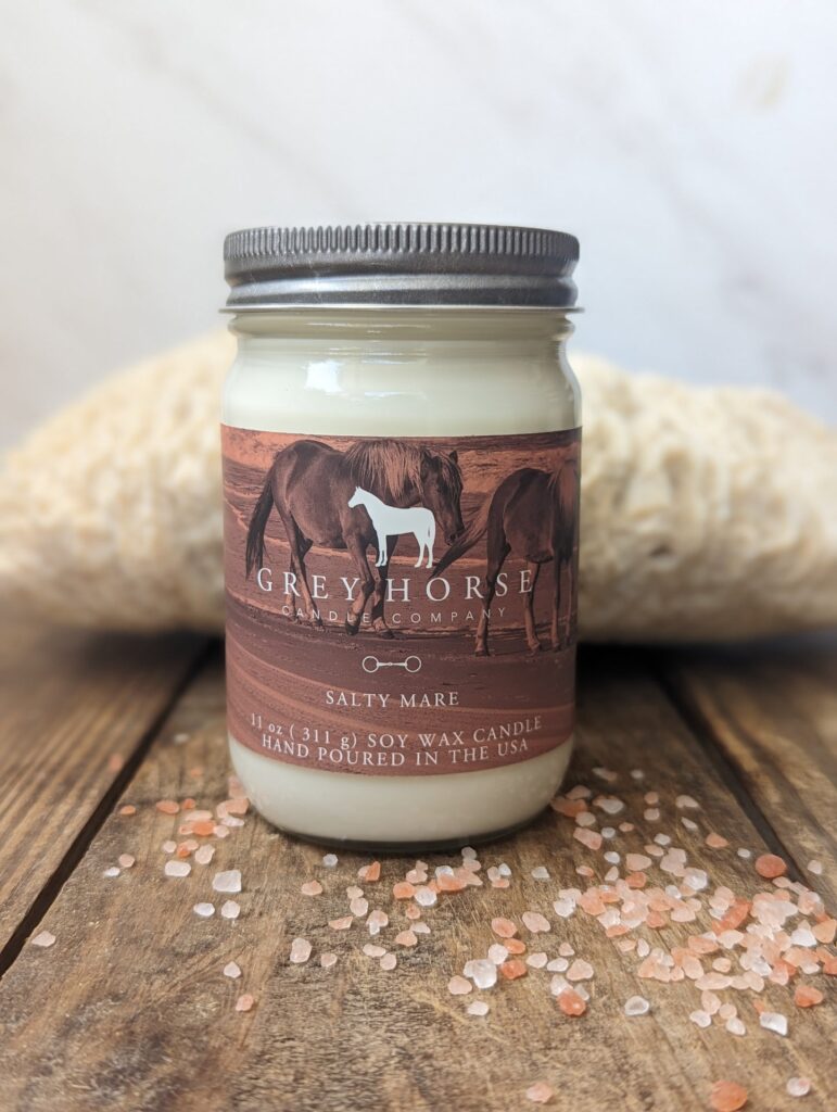 The Salty Mare Jar candle, featuring the Grey Horse Candle Company label, rests on a wooden surface amidst scattered pink salt crystals, set against a textured light background.