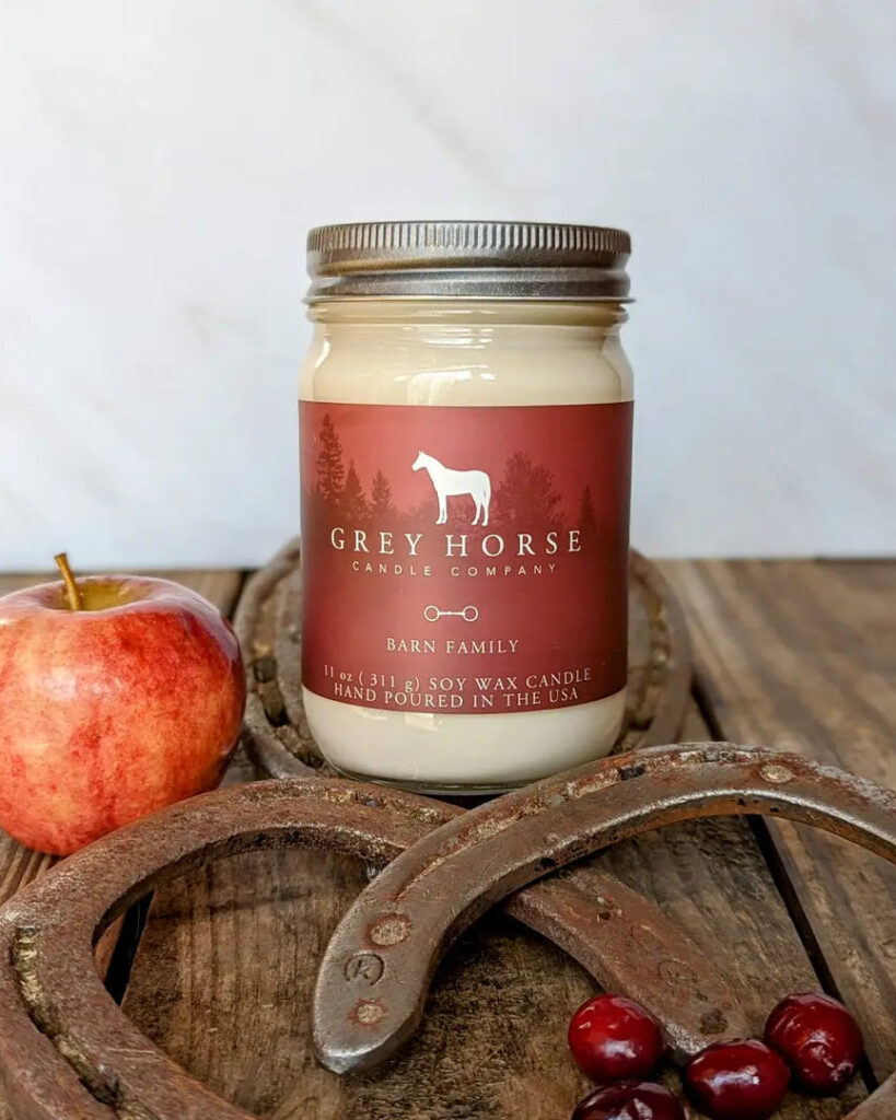 A Barn Family Jar candle from Grey Horse Candle Company rests on a wooden surface, surrounded by an apple, cranberries, and two horseshoes. The label reads Barn Family, beautifully evoking the warm and inviting feeling of a cosy barn gathering.