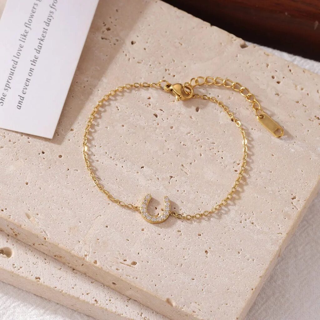 A delicate gold bracelet features a small, horseshoe-shaped charm encrusted with tiny clear stones, adding a subtle touch of elegance and sparkle to this beautiful horseshoe bracelet. Displayed on a textured beige surface next to a piece of white paper, it’s the perfect accessory for any occasion.
