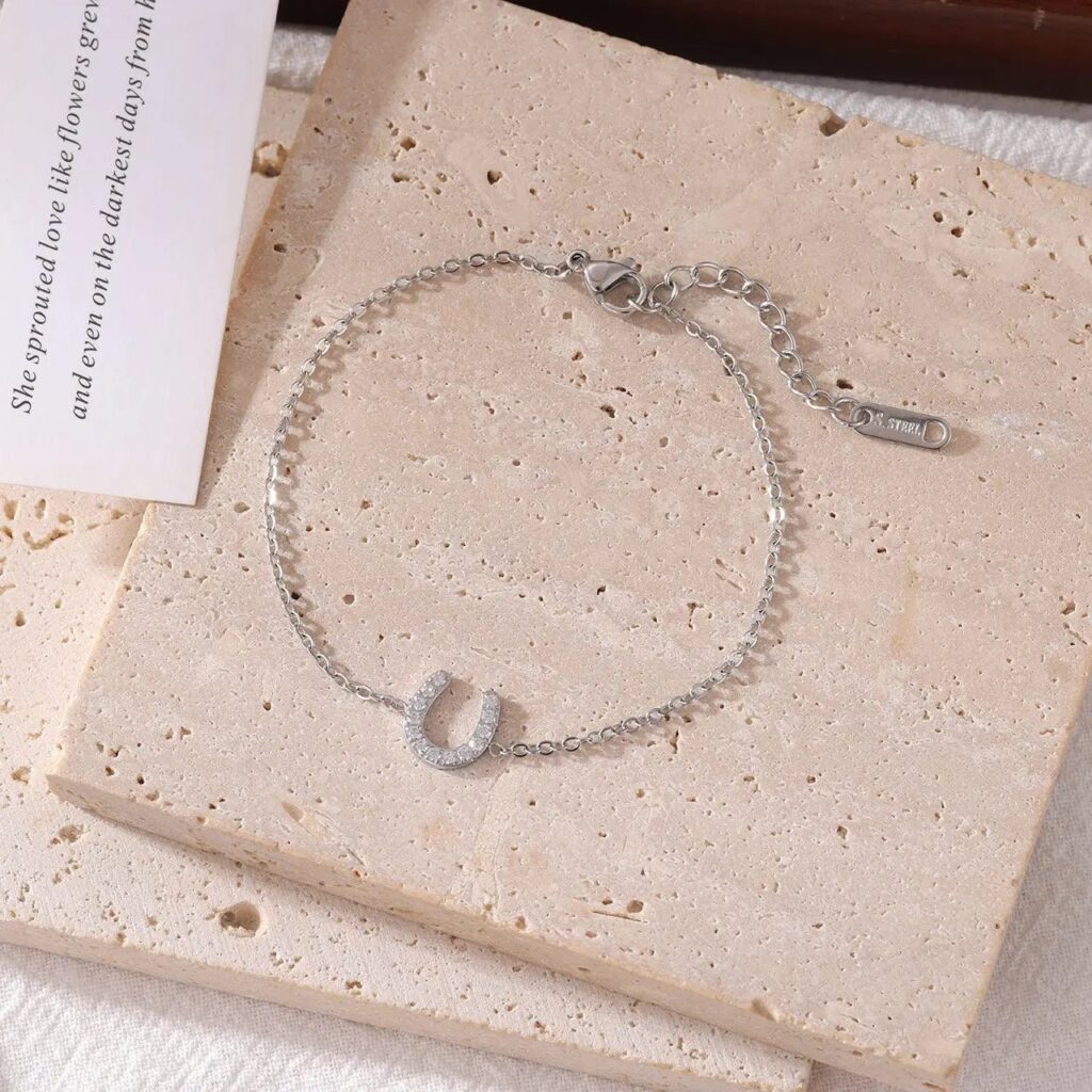 A delicate silver chain bracelet with a crystal-studded horseshoe charm is displayed on textured beige stone tiles. A partially visible card with cursive writing is placed next to the bracelet.