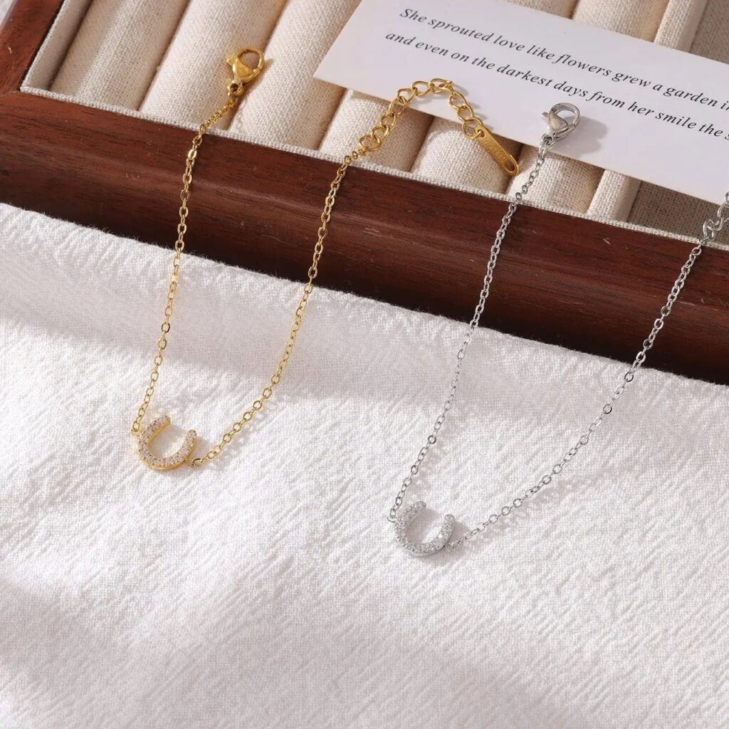 Two delicate chain bracelets, one gold and one silver, each featuring a horseshoe charm, rest on a white textured fabric. A wooden box with cream fabric slots and a partially visible note are in the background.