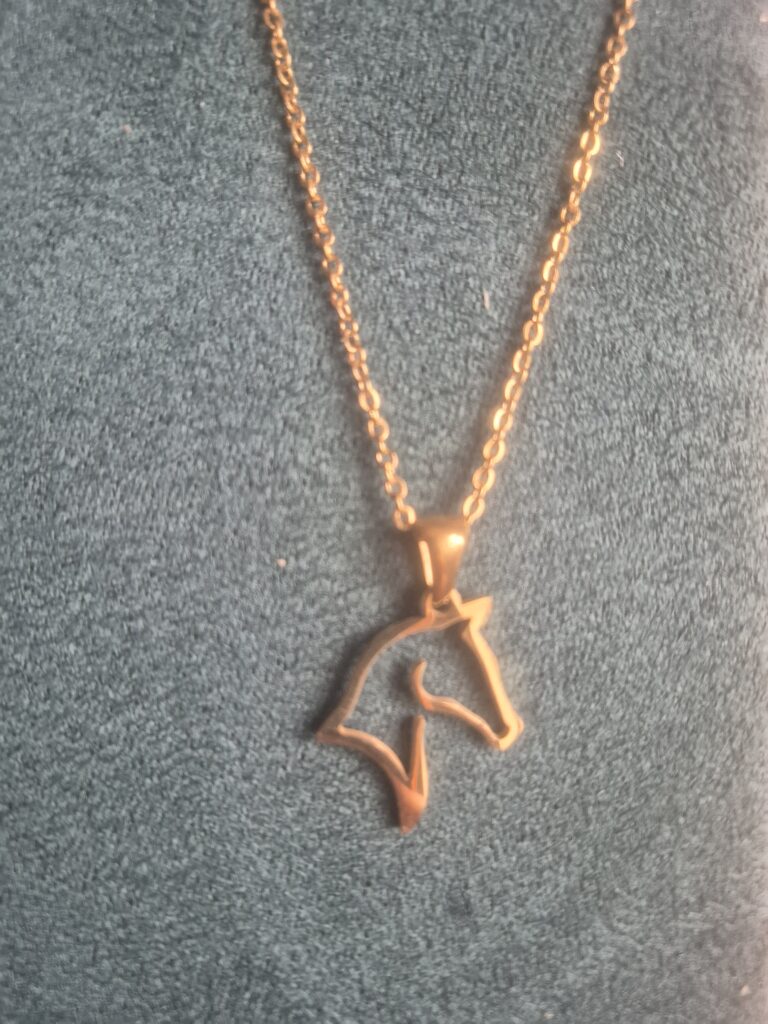 A gold necklace with a pendant shaped like a minimalist horse head outline, displayed on a textured dark green background.