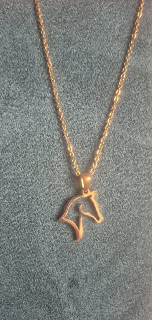 A gold necklace with a pendant shaped like a horse's head outline lies on a textured dark green surface.