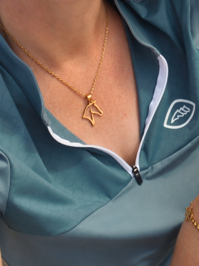 A person wearing a teal and white zip-up top with a geometric pattern and a gold horse head necklace. Part of a gold bracelet is also visible on their wrist.
