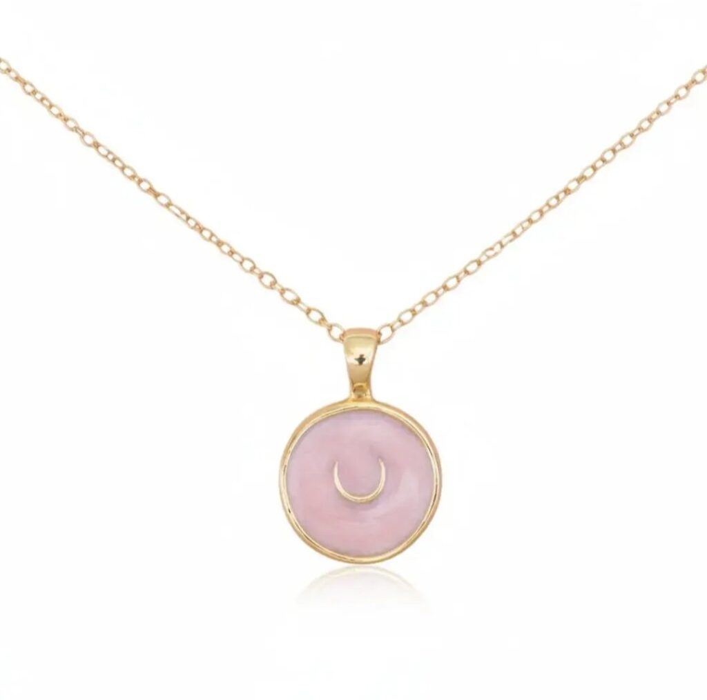 A gold chain necklace with a round pink pendant featuring a small gold crescent moon in the centre, displayed on a white background.
