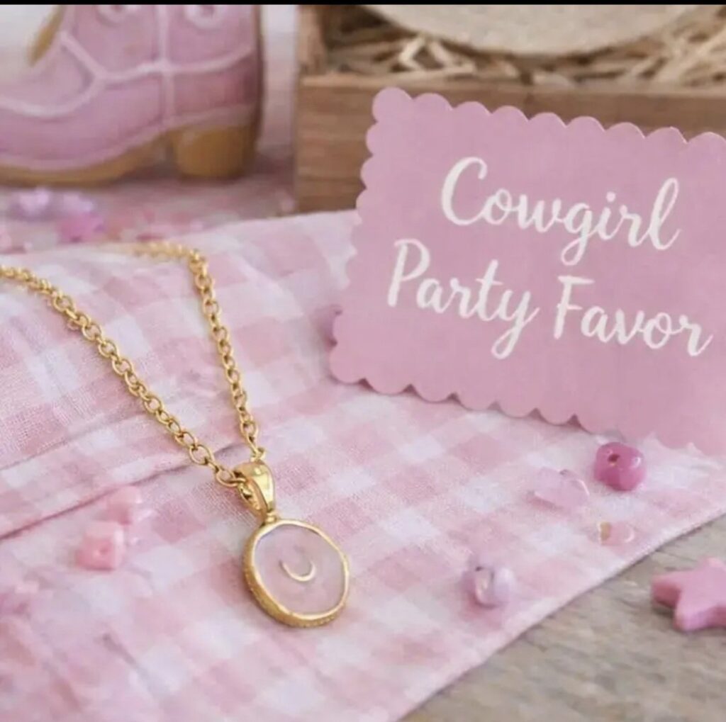 A gold necklace with a crescent moon pendant rests on a pink checked cloth, surrounded by beads. A pink card nearby reads Cowgirl Party Favour in white script.