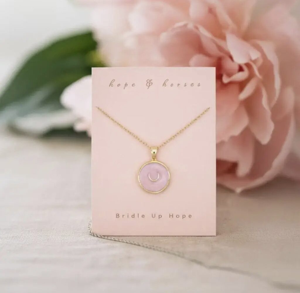 A gold necklace with a round pink pendant on a card reading hope & horses and BRIDLE UP HOPE, set against a soft background with a large pink flower.