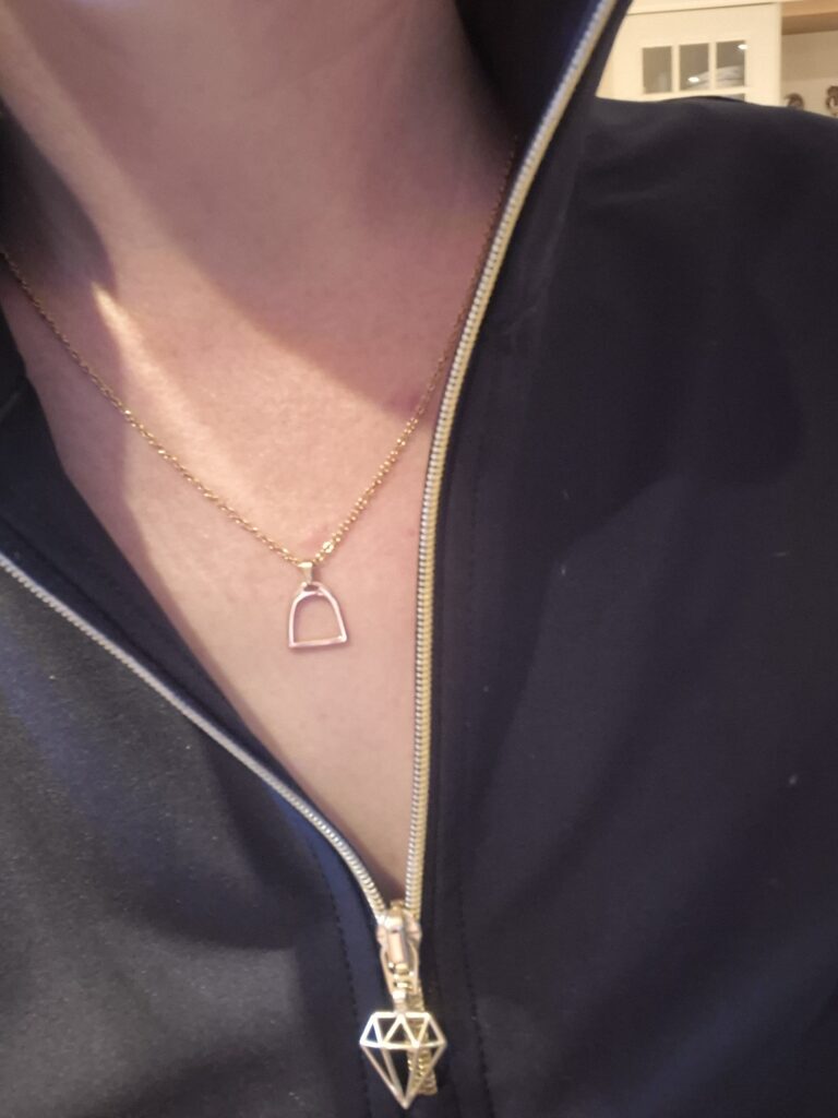 A person wearing a gold necklace with a simple geometric pendant, partially visible inside a dark zip-up jacket with a diamond-shaped zip pull.