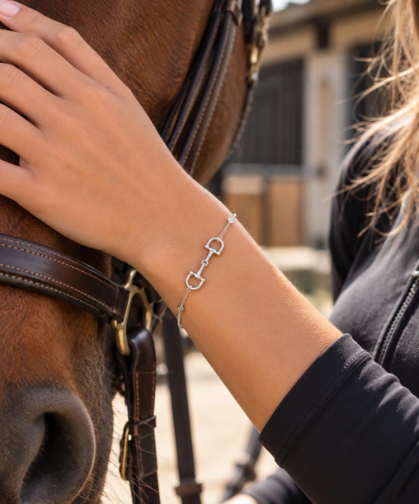 A person gently touches a horse's face, showcasing a silver bracelet with equestrian-inspired charms. The horse is wearing a bridle and the background appears to be a stable.